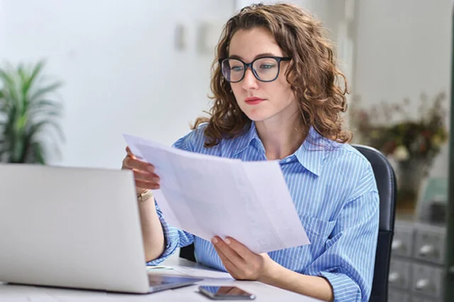 A woman with glasses sits at a desk, reviewing documents while working on a laptop