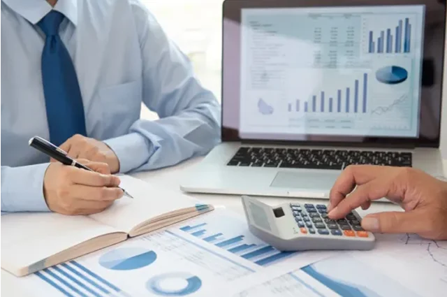 reviewing financial charts and data at a desk, writing in a notebook, using calculator