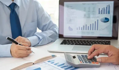 reviewing financial charts and data at a desk, writing in a notebook, using calculator