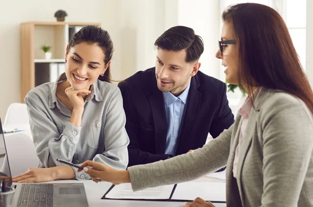 woman in glasses points to a laptop screen while a couple looks on attentively in an office setting