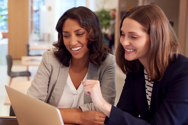 women smiling and looking at a laptop together in a modern office space, suggesting collaboration