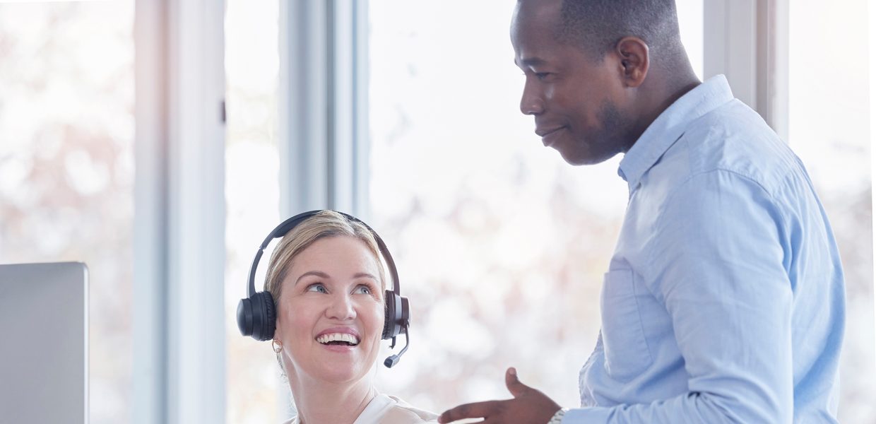 A smiling assistant wearing a headset talks with a colleague at her desk